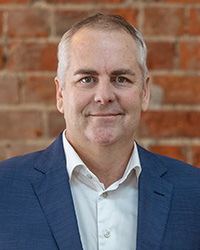 A smiling man in a navy suit standing in front of a red-brick wall.