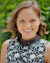 A headshot of a smiling woman with short, brown hair standing in front of a green, leafy backdrop.