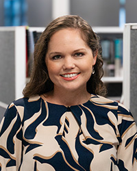 A headshot of a smiling woman with wavy, brown hair standing in an office building.